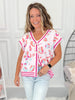 Woman wearing a colorful patterned shirt and light jeans in a room with a white wall and decorative items.